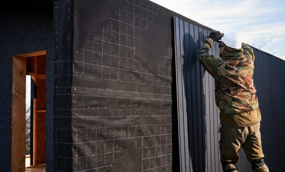 Male builder installing black corrugated iron sheet used as facade of future cottage. Man worker building wooden frame house. Carpentry and construction concept.