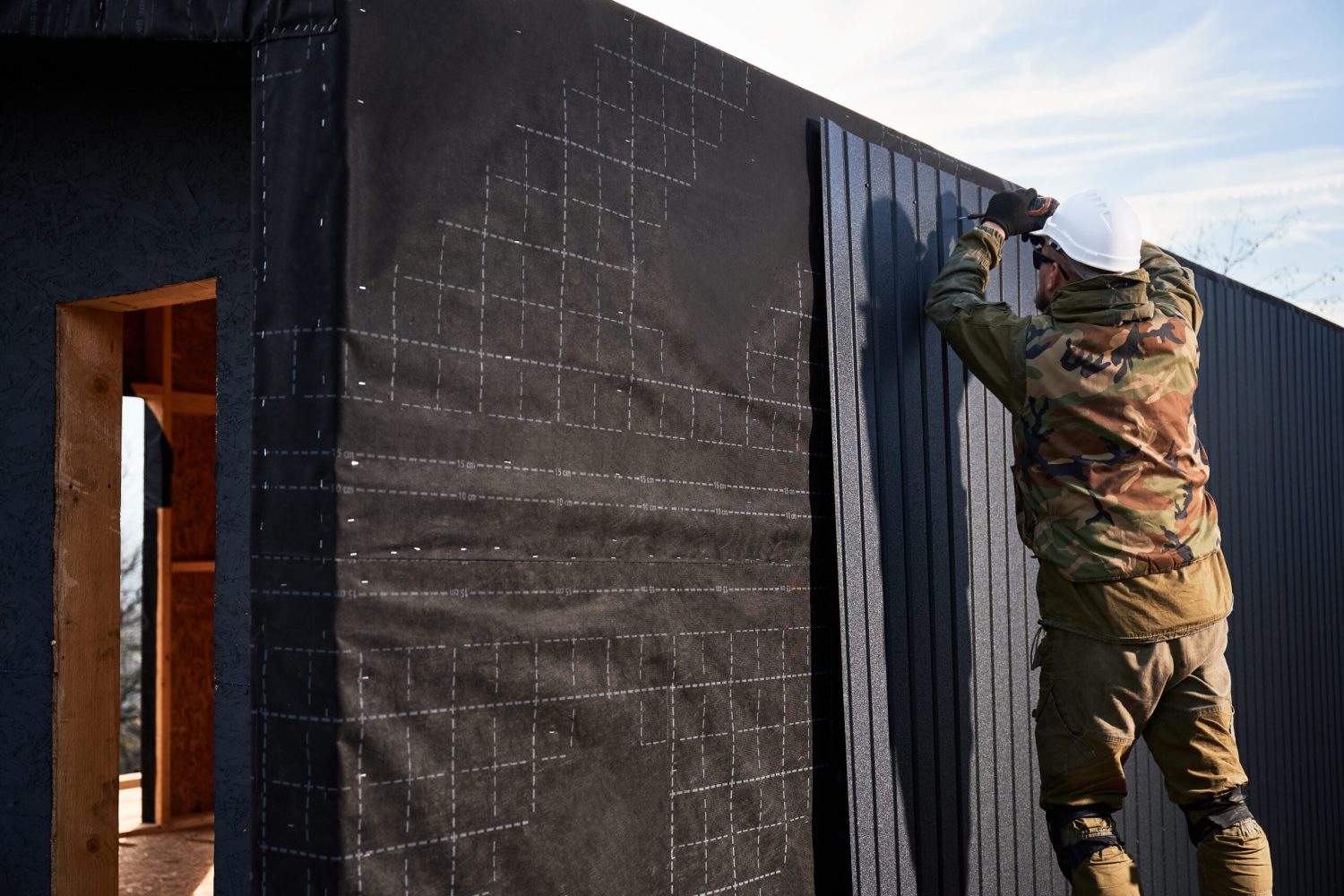 Male builder installing black corrugated iron sheet used as facade of future cottage. Man worker building wooden frame house. Carpentry and construction concept.
