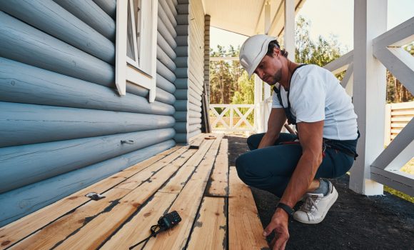 Construction worker sitting on his haunches on porch of building while placing wooden boards on floor